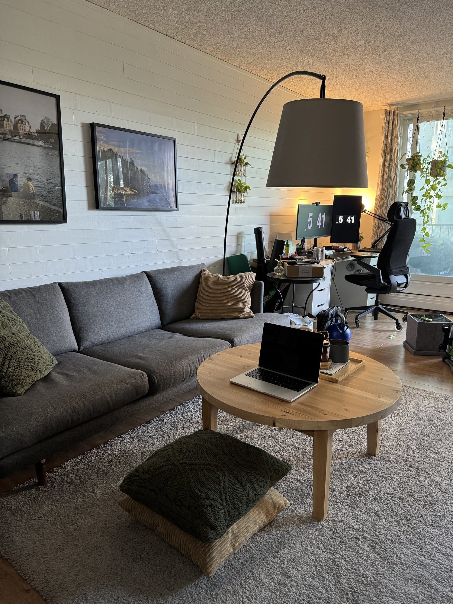 A living room with a grey sofa, a wooden coffee table with a laptop, stacked floor cushions, and a workspace in the background with dual monitors and a black office chair