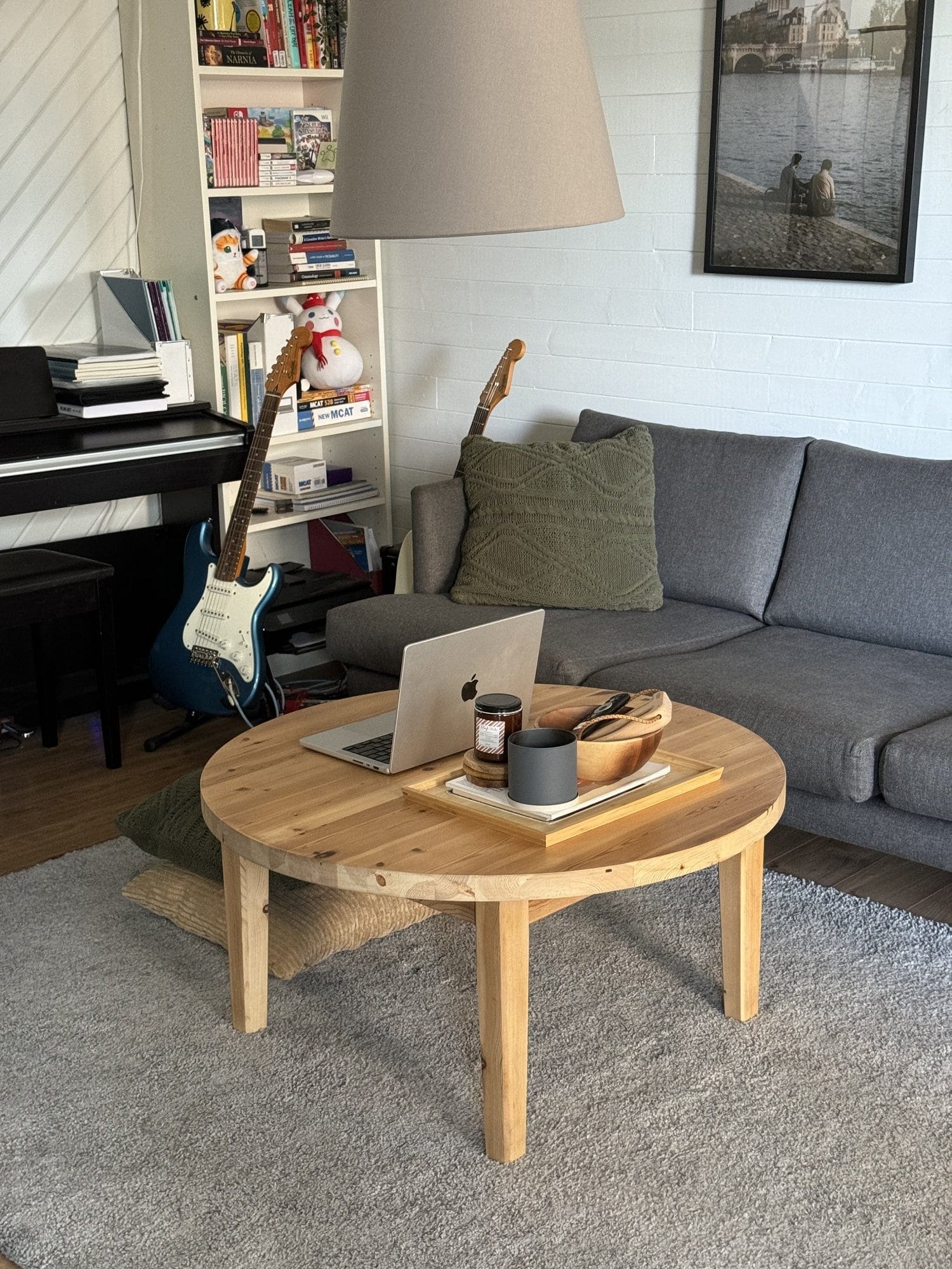 A living room with a wooden coffee table holding a laptop and tray, a grey sofa with green cushions, a bookshelf, a piano, and two guitars leaning against the furniture