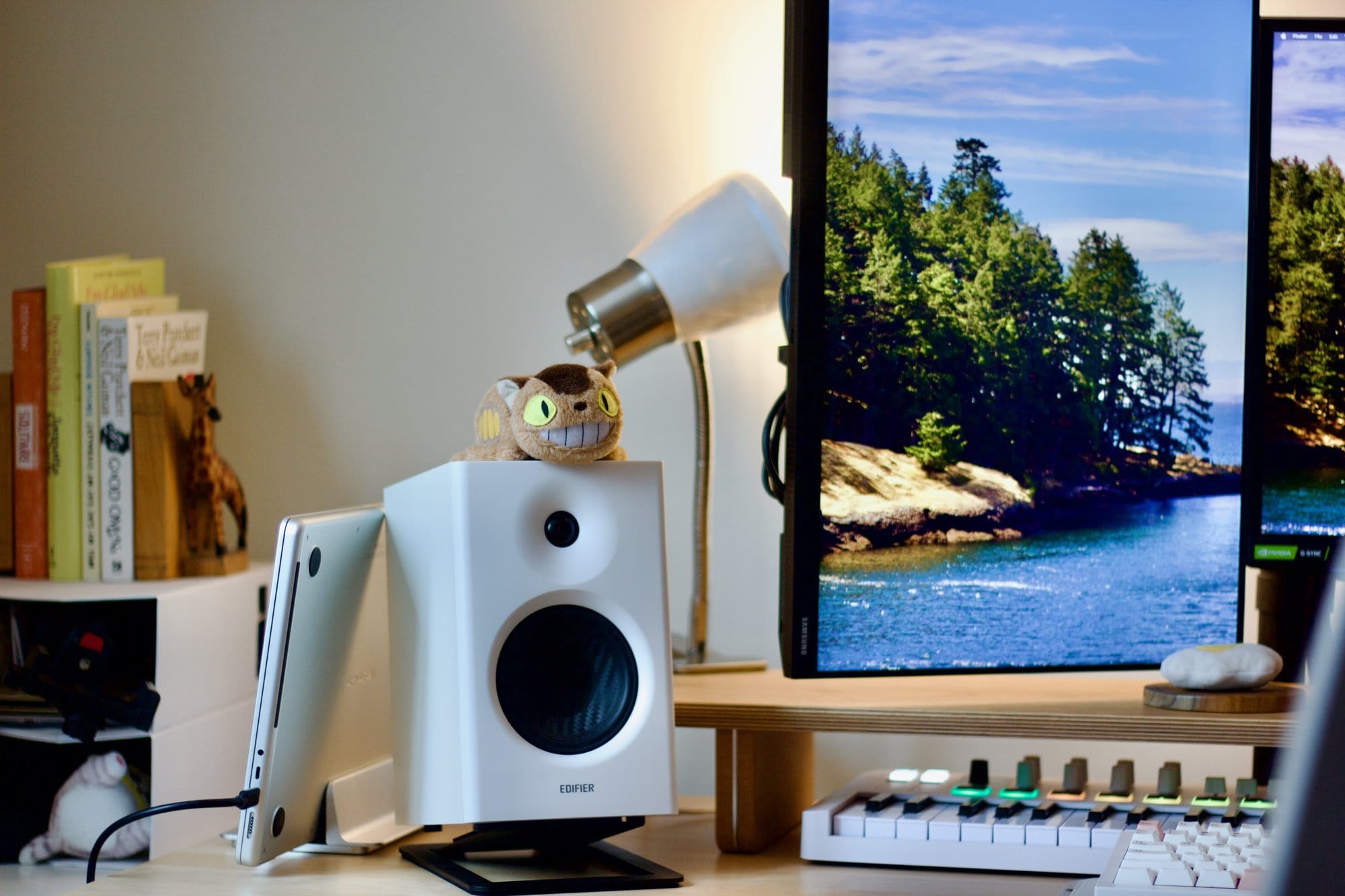 A workspace setup with a Samsung 24″ FHD vertical monitor, an Edifier MR4 speaker with a Catbus plush on top, a MacBook in a vertical stand, an Arturia MiniLab 3 MIDI keyboard, and a bookshelf with novels