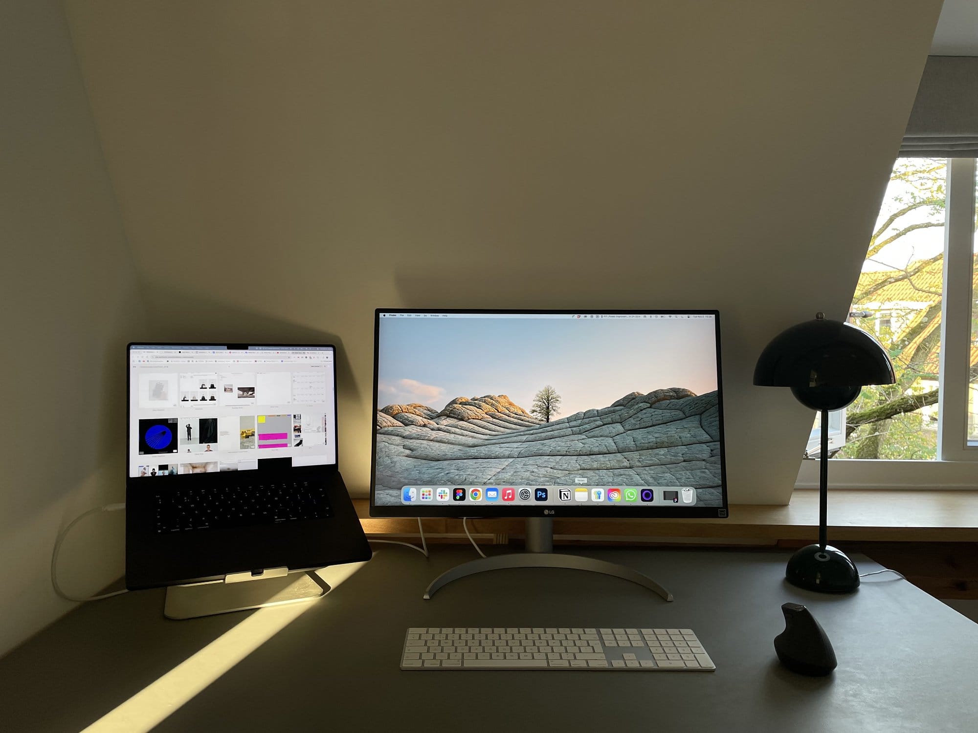 A workspace setup with an LG UltraFine 27UN850-W monitor, a MacBook Pro 14″ on a raindesign mStand laptop stand, an Apple wireless keyboard with numpad, a Logitech MX Master 2 mouse, and a black table lamp near a window