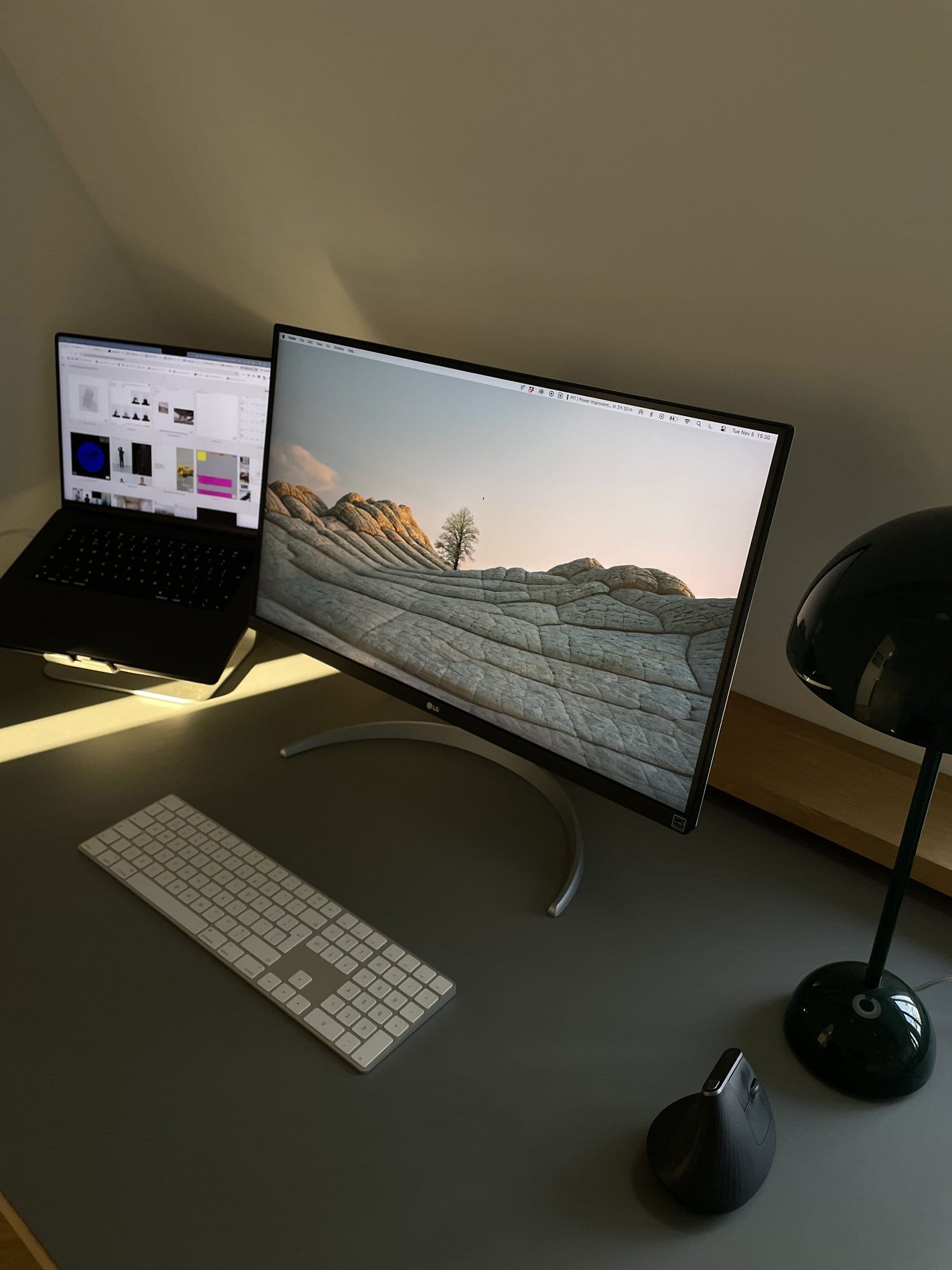 An angle view of a workspace setup with an LG UltraFine 27UN850-W monitor, a MacBook Pro on a laptop stand, an Apple wireless keyboard, a Logitech MX Master 2 vertical mouse, and a black table lamp on a pale green desk with sunlight casting shadows