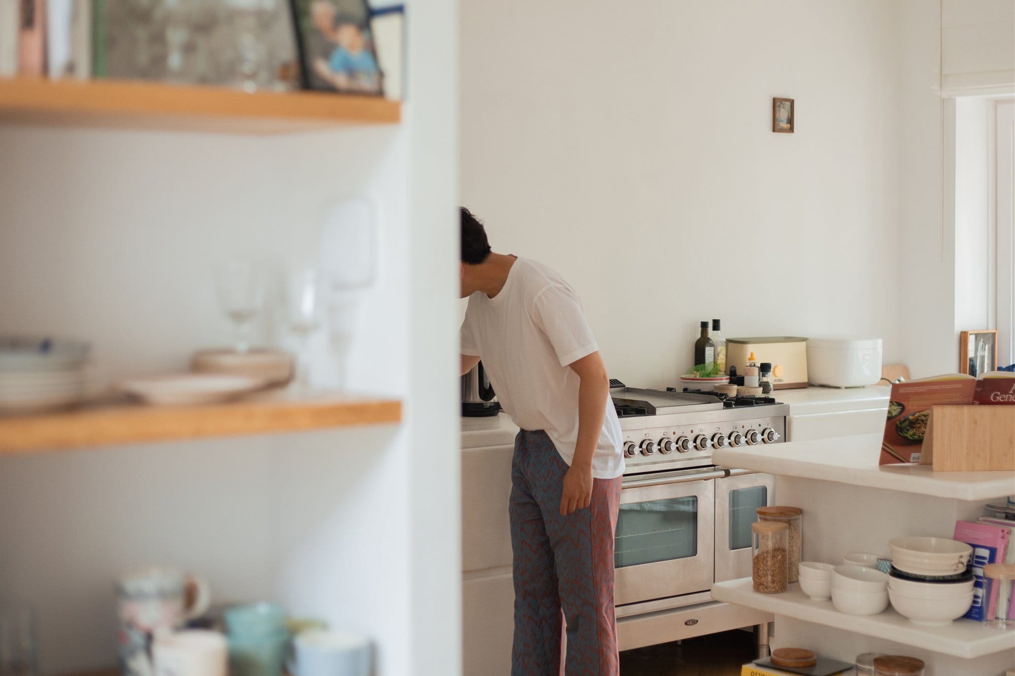 A kitchen scene with open wooden shelves holding dishes and glassware, while a person in a white t-shirt and patterned trousers leans over the counter near a gas stove