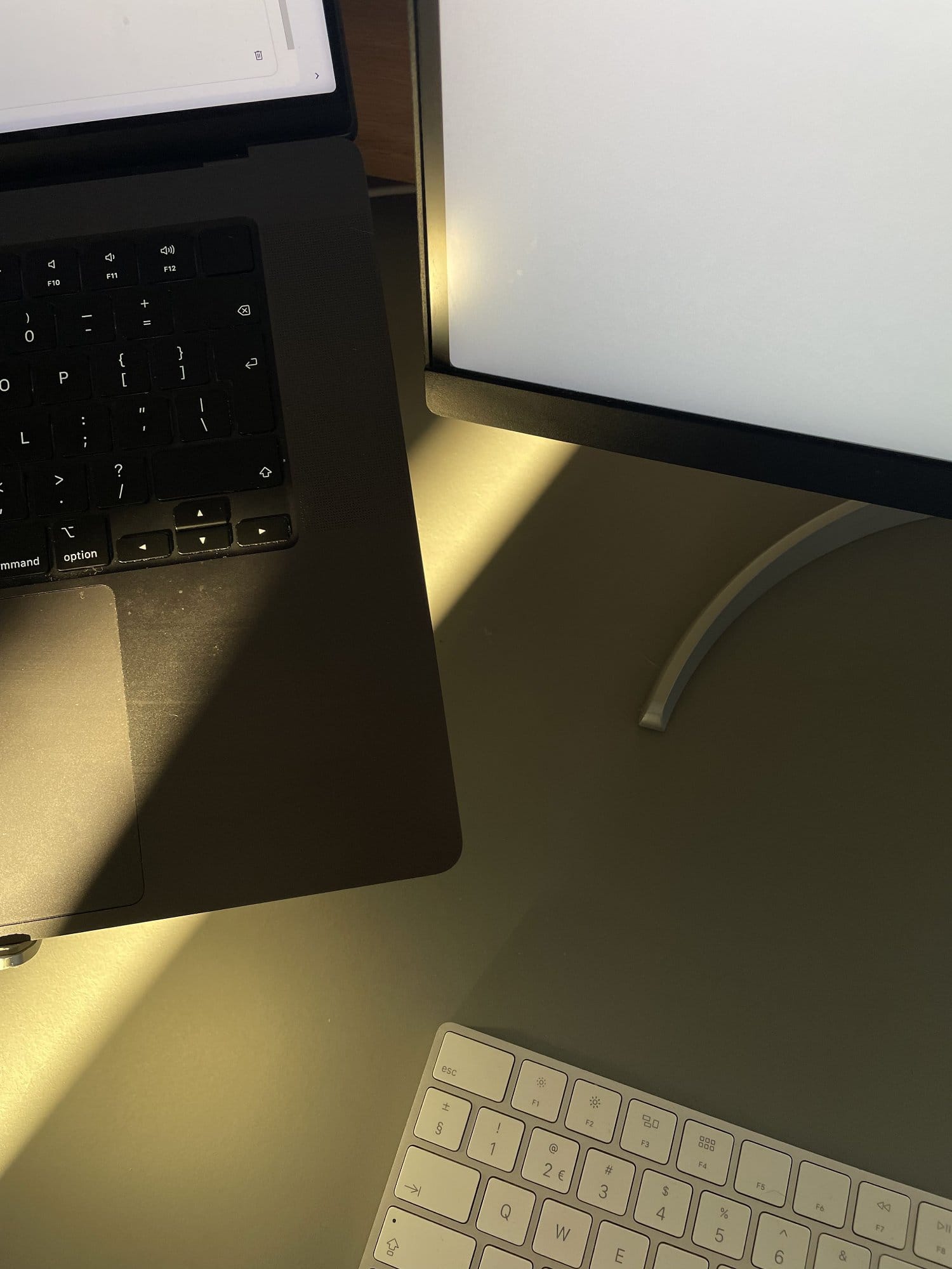 A close-up of a workspace setup with a MacBook Pro laptop, an LG UltraFine 27UN850-W monitor, and an Apple keyboard on a dark green desk, with sunlight casting angular shadows