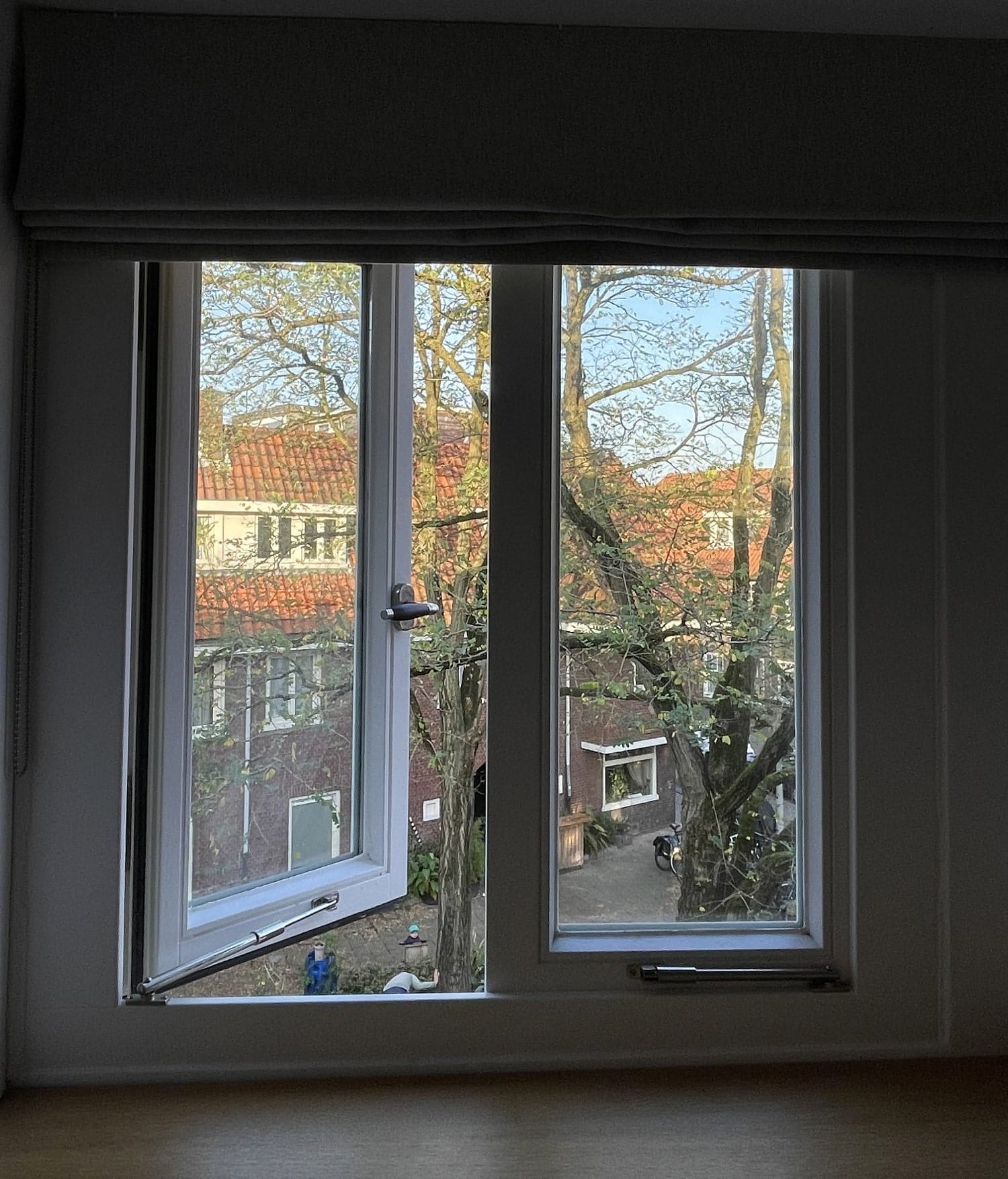 An open window with a grey fabric blind, showing a view of red-roofed buildings, trees with budding leaves, and a small courtyard with plants