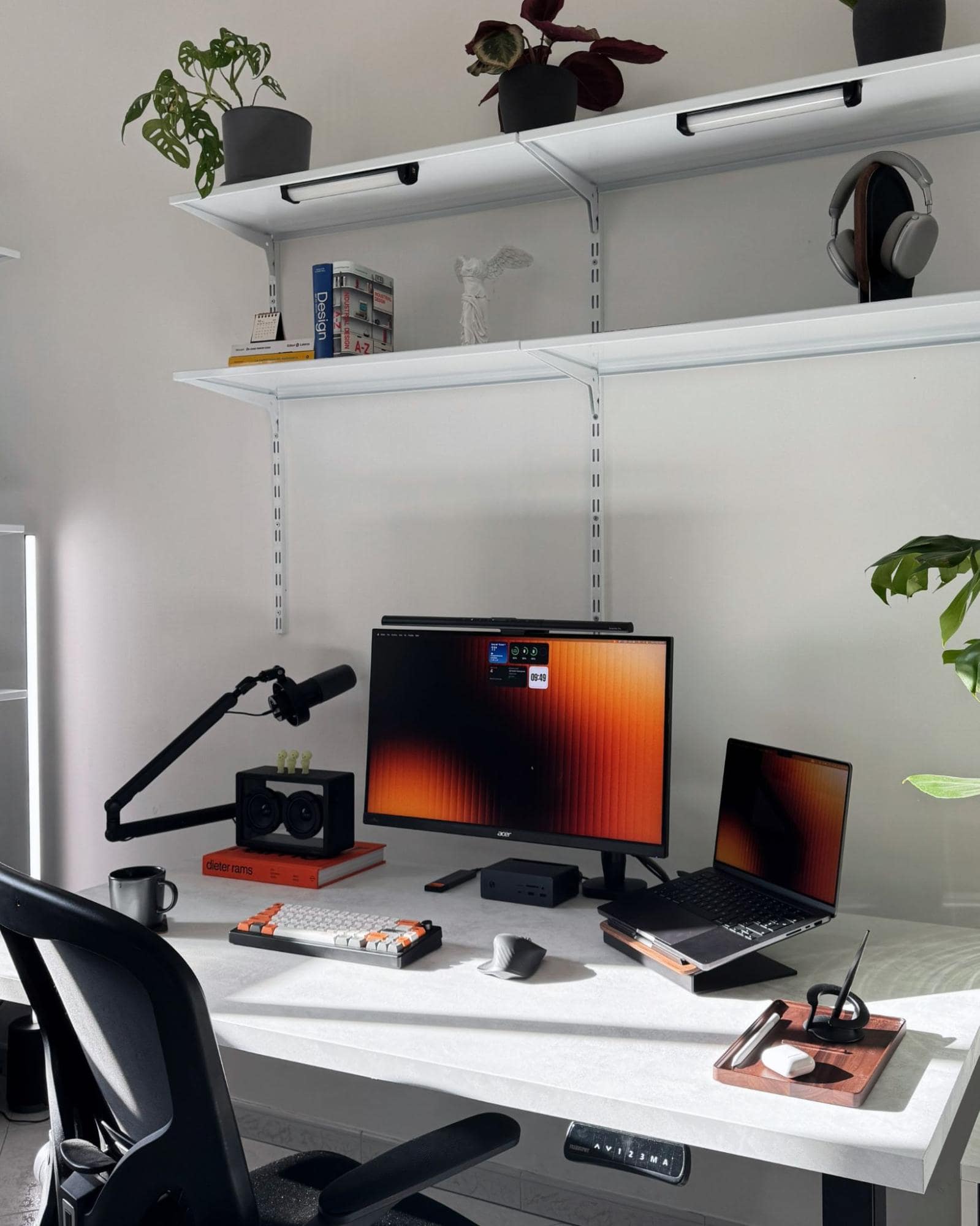 A desk setup shot from a straight-on angle, showing an Acer XV272UP monitor, a MacBook Pro 14″ M3 Pro on a laptop stand, and a FIFINE K688 microphone on an adjustable arm, with a Keychron K3 Max keyboard and Logitech MX Master 3S mouse on the white desk, while shelves above hold plants, books, and headphones