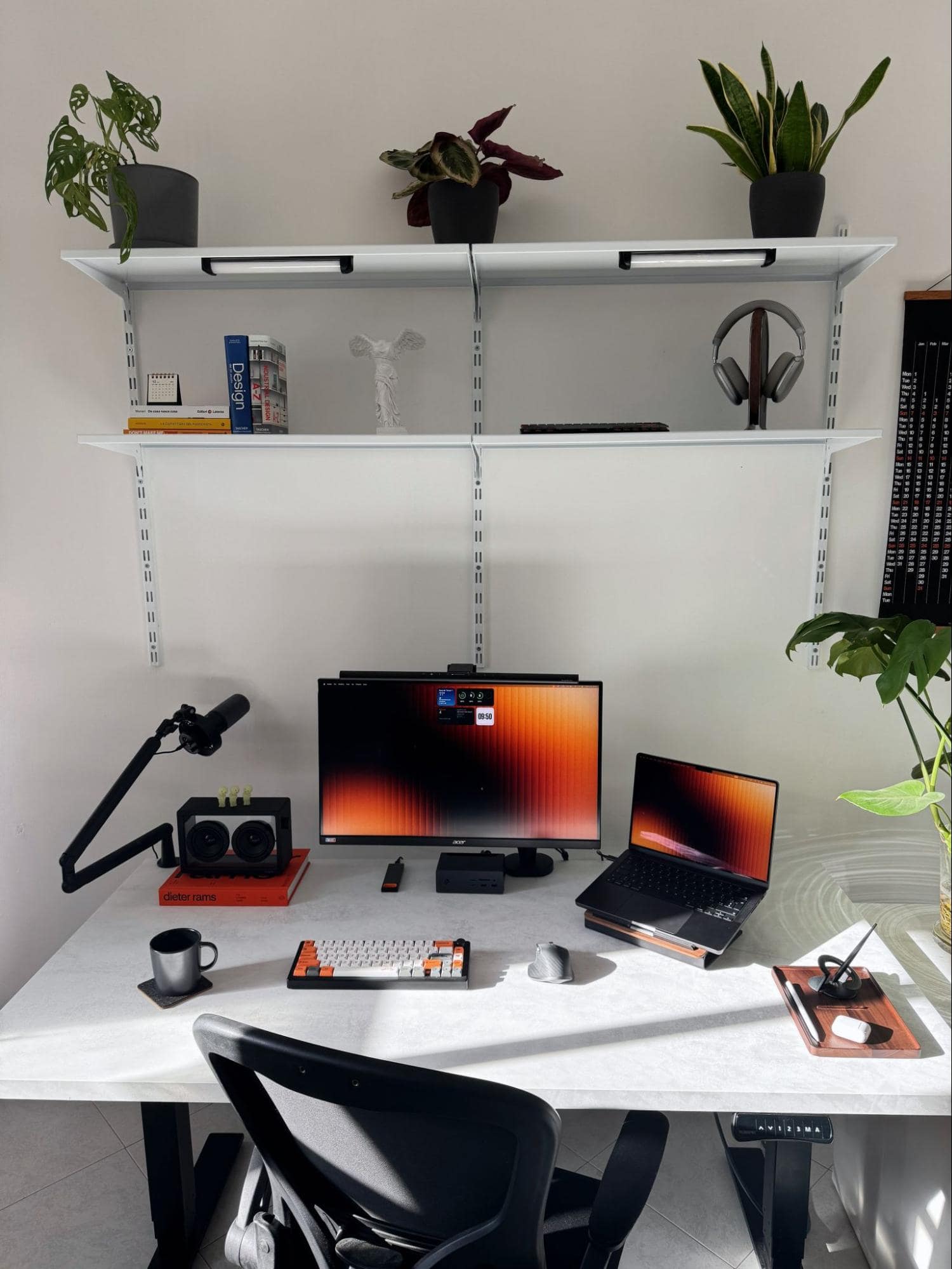 A workspace setup with an Acer XV272UP monitor, a MacBook Pro 14″ M3 Pro on a laptop stand, and a Keychron K3 Max keyboard, arranged on a white desk with a microphone, speakers, and shelves holding plants, books, and headphones