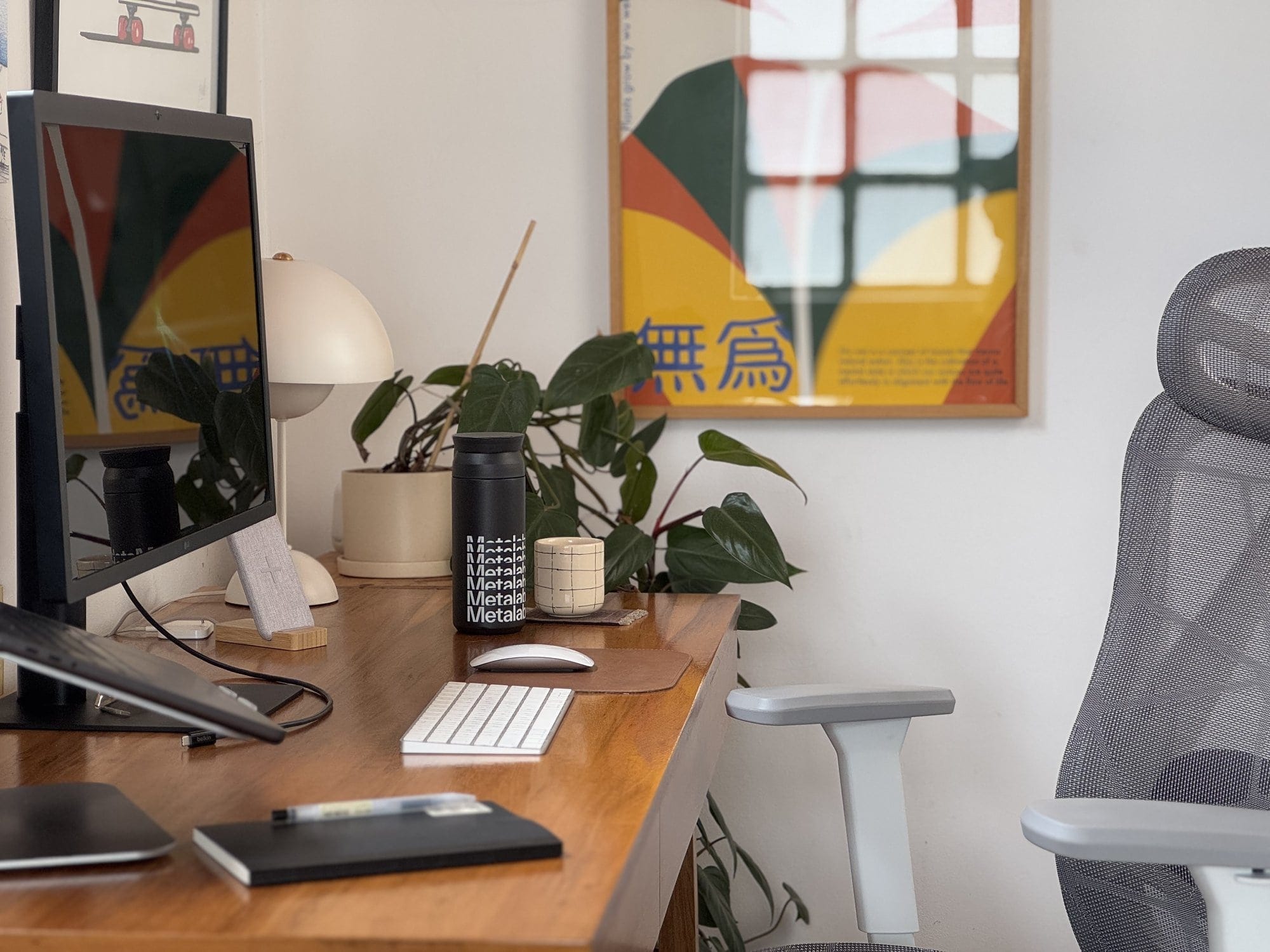 A workspace with an LG UltraFine 4K monitor, an Apple Keyboard, an Apple Magic Mouse, an Actek Floe Plus EC727 chair, with a notebook, a water bottle, and a potted plant on a wooden desk