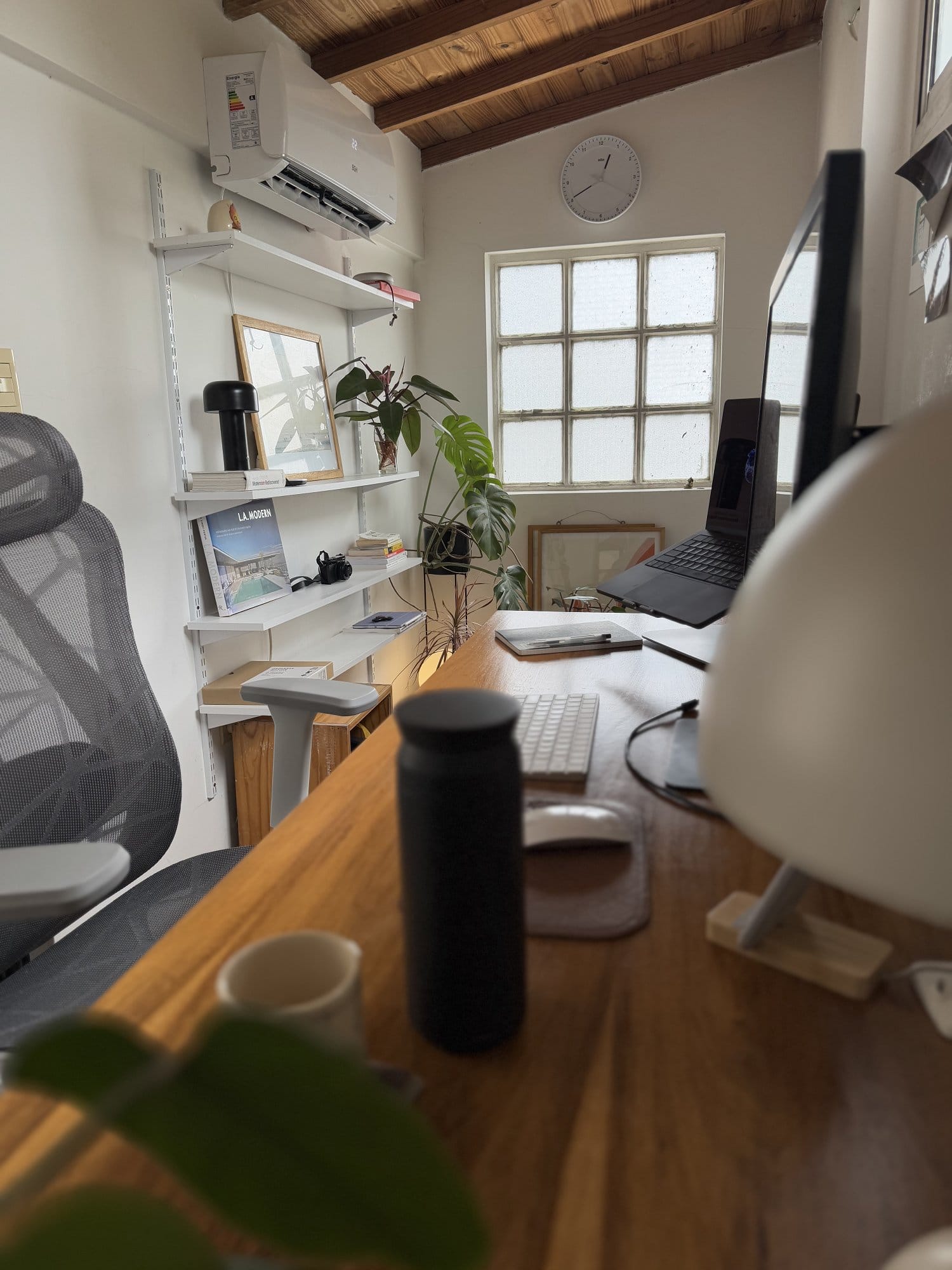 A small home office desk setup with an Apple MacBook Pro M3 Pro on a Rain Design mStand360, an external monitor, an Apple Keyboard, an Apple Magic Mouse, and an Actek Floe Plus EC727 chair, with shelves and a window in the background