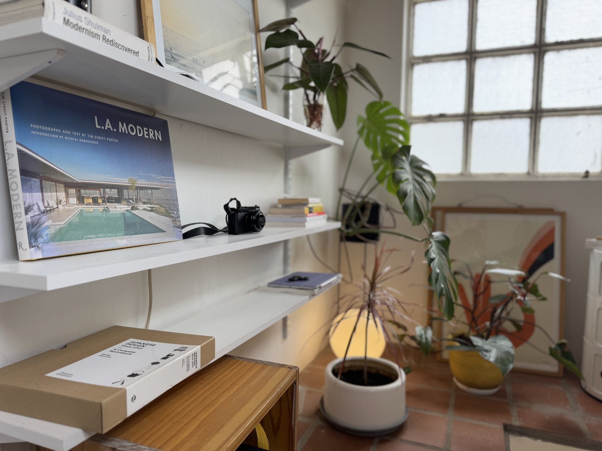 White wall-mounted shelves with books, a black camera, and some potted plants