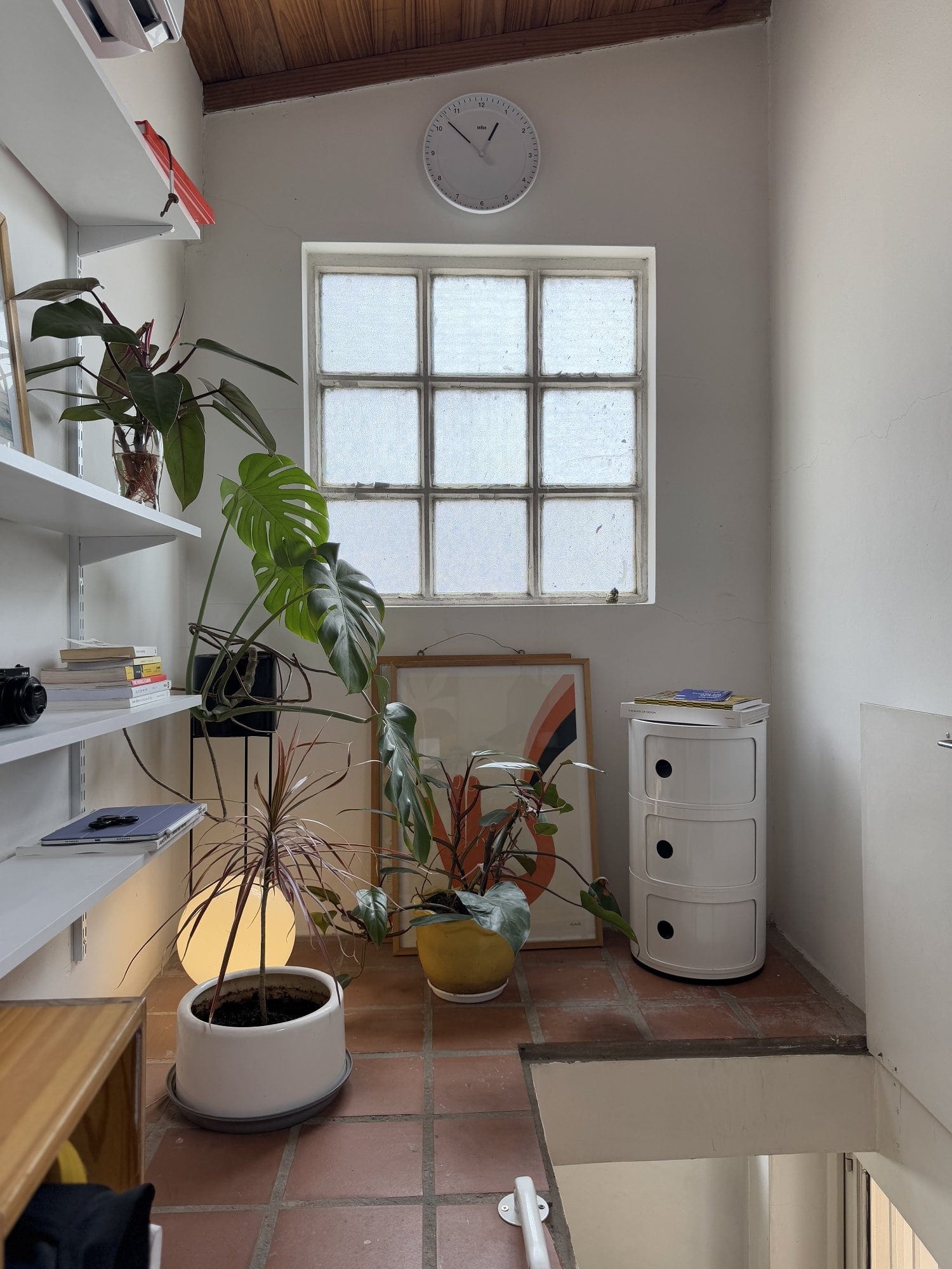 A small nook with a white Kartell Componibili storage unit, a mix of potted plants, a framed abstract print leaning against the wall, and shelves holding books and a camera, illuminated by natural light from a window