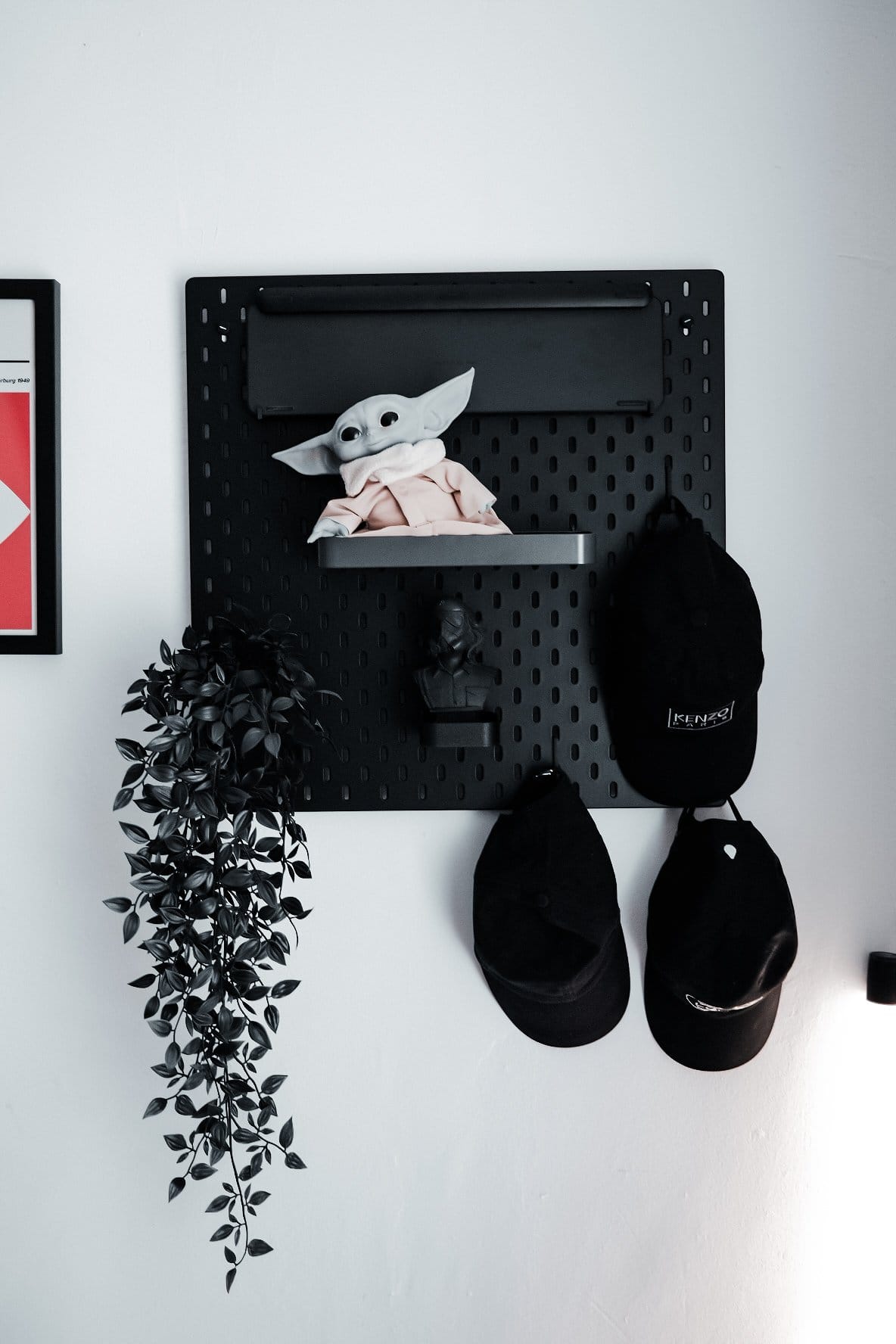 A black pegboard mounted on the wall, holding a Grogu figure, a small bust sculpture, a hanging plant, and several black caps, next to a framed Helvetica poster