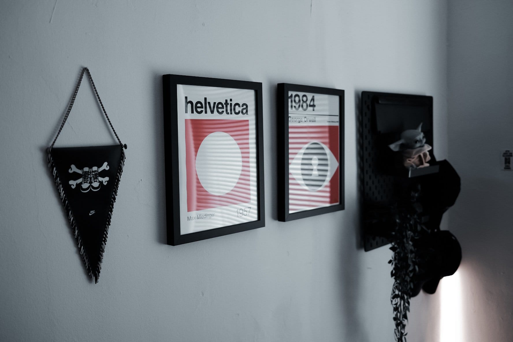 A wall setup with framed Helvetica and 1984 posters, a Nike pennant, and a pegboard holding caps, a hanging plant, and a small Grogu figure on a shelf