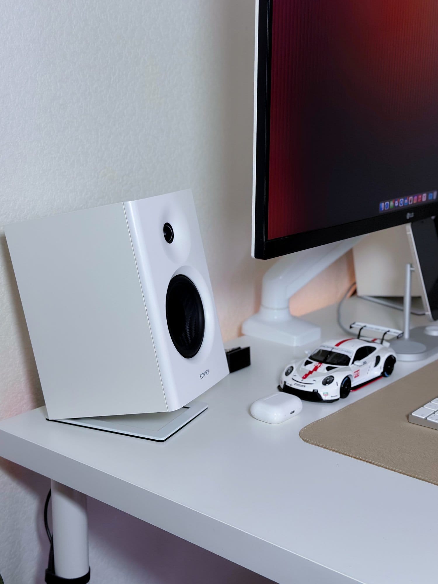 A close-up of a workspace setup with an Edifier MR4 speaker, an LG 27UL650-W monitor, a Porsche model car, and a beige desk mat on a white desk