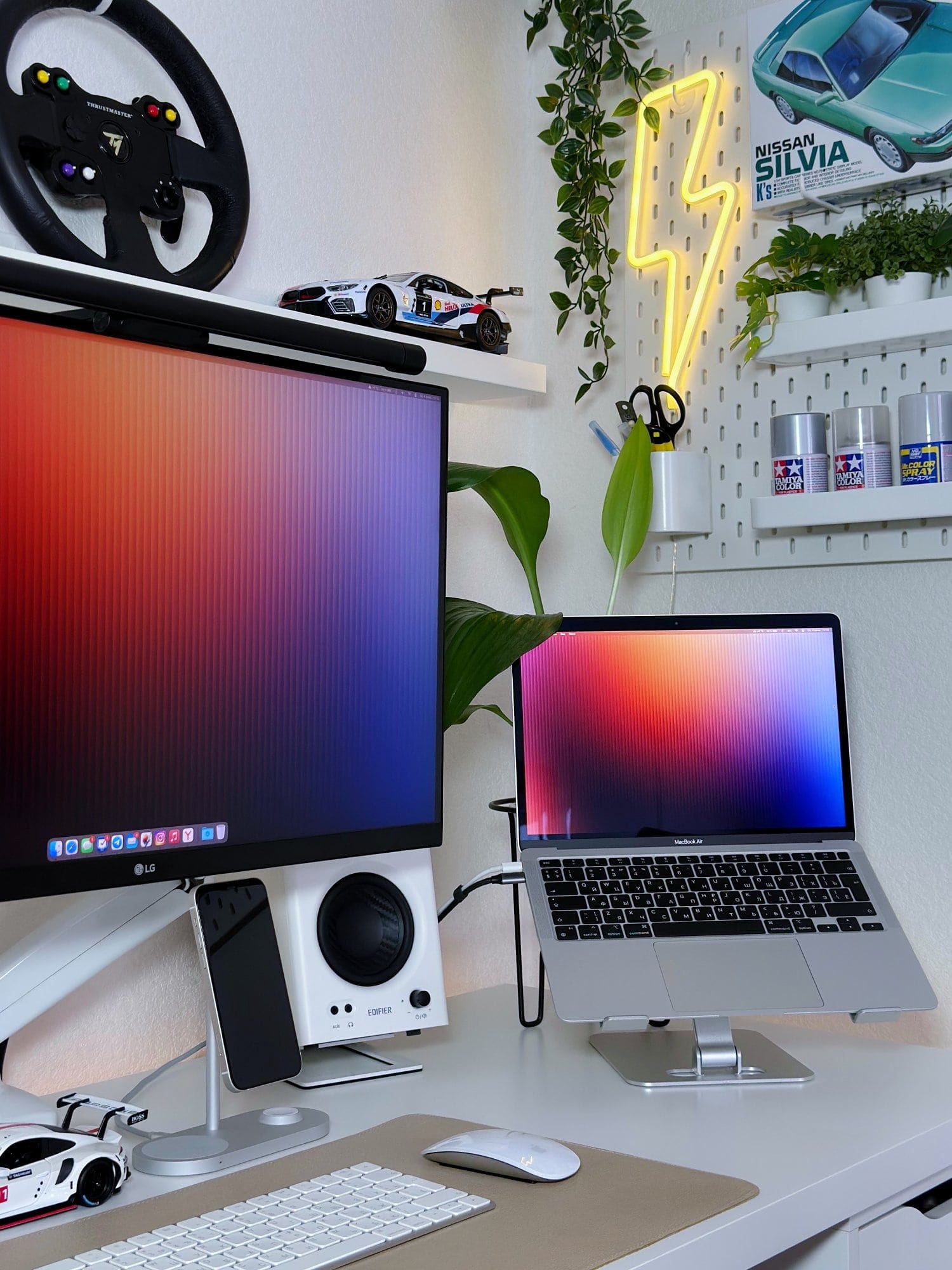 A desk setup with an LG 27UL650-W monitor, a MacBook Air M1 2020 on an aluminium stand, Edifier MR4 speakers, and a neon lightning bolt sign on a pegboard with plants and model car accessories