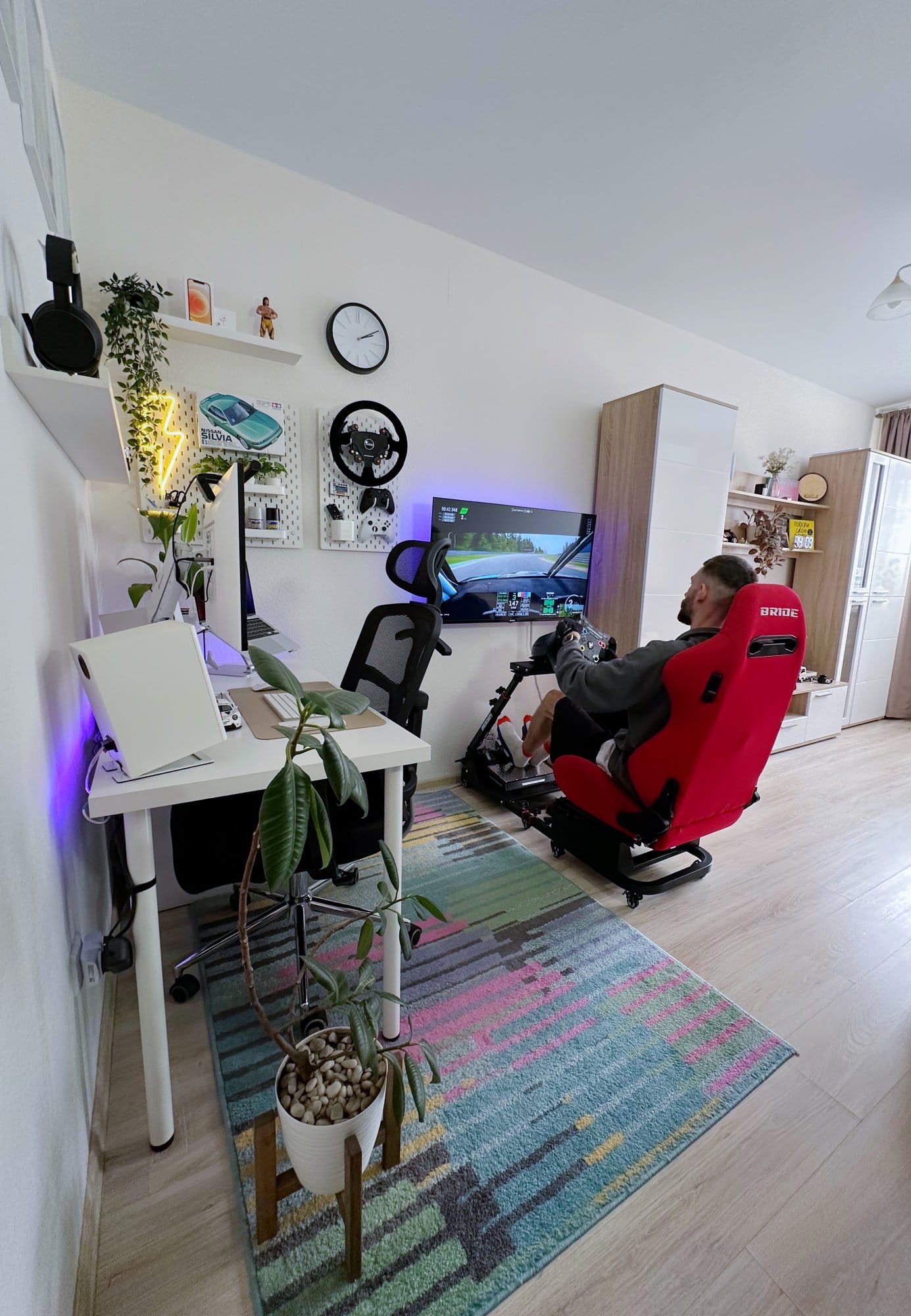 A workspace setup with an LG 27UL650-W monitor, a MacBook Air M1 2020, and Edifier MR4 speakers on a white desk, with a TetChair MESH-6 office chair nearby, while a person is using a racing simulator with a BRIDE red racing seat