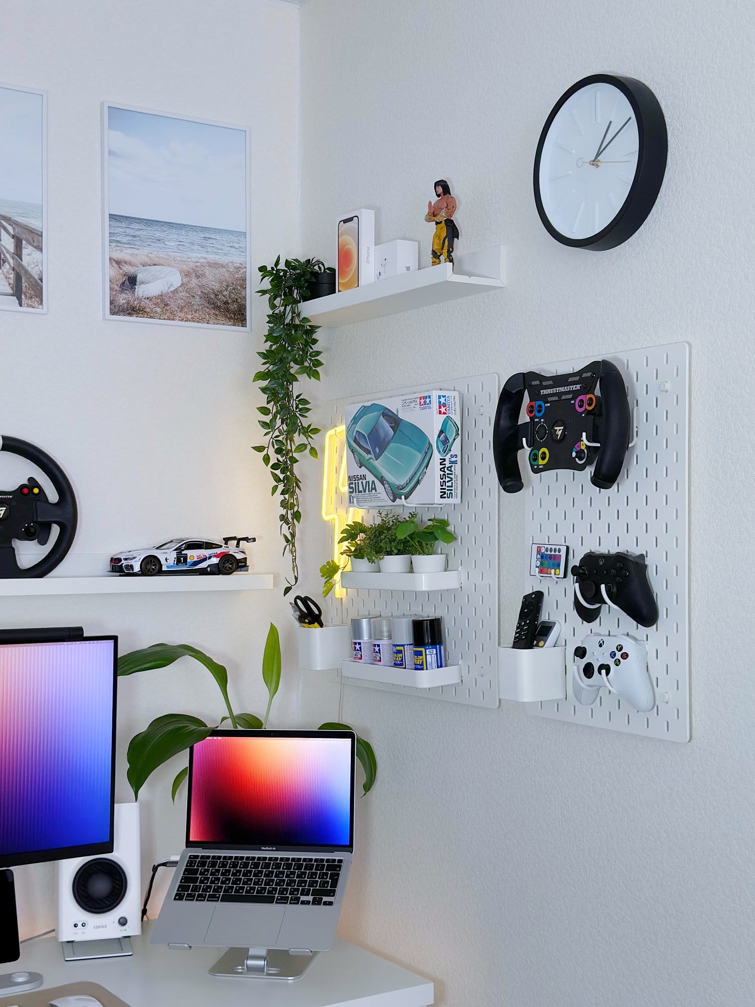 A corner of a minimal workspace setup with a MacBook Air M1 2020 on an aluminium stand, Edifier MR4 speakers, a pegboard holding gaming controllers and accessories, and wall shelves with plants, a clock, and a car model