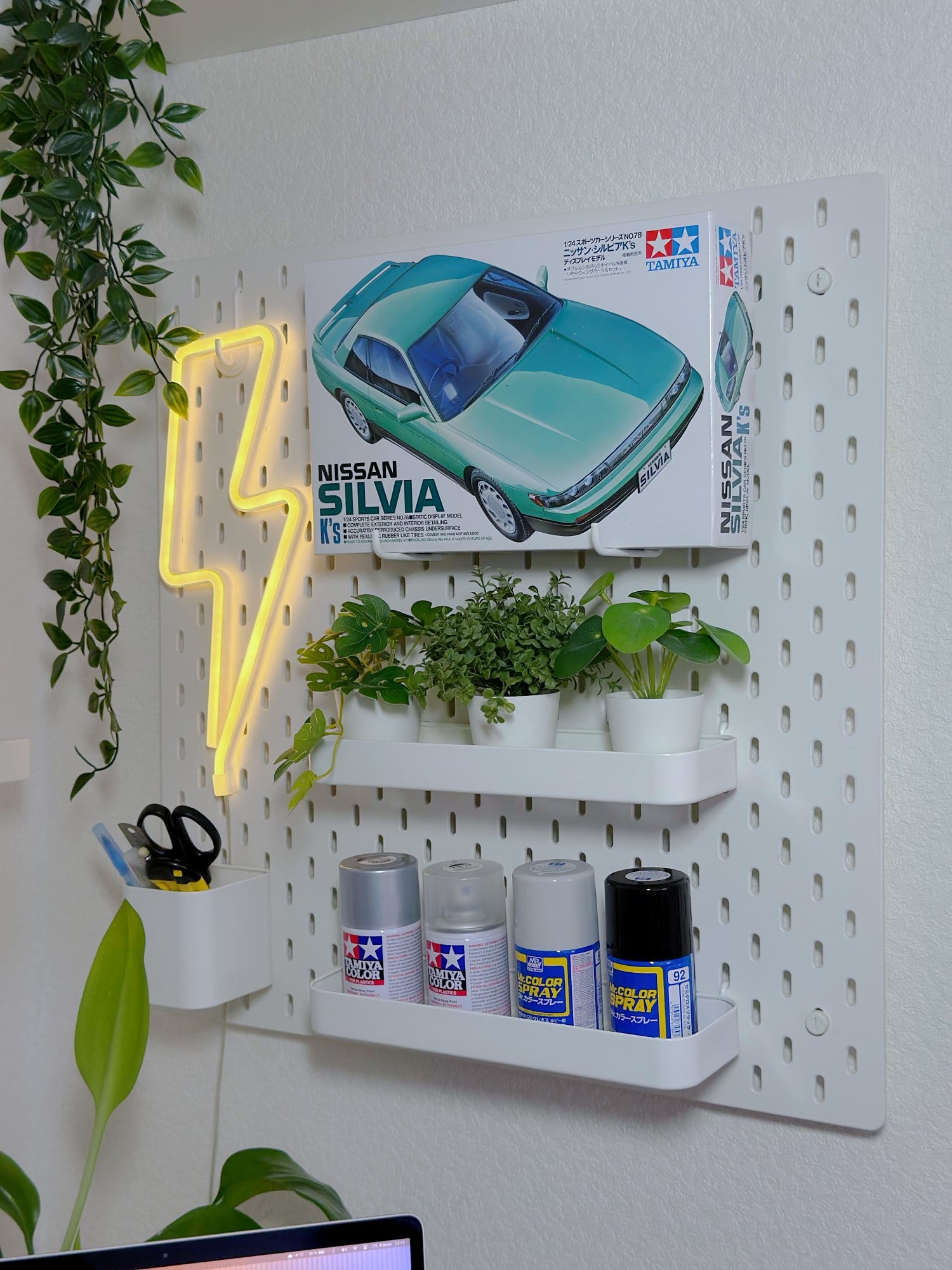 A pegboard with a Tamiya Nissan Silvia K’s model kit, spray paints, small potted plants, a neon lightning bolt sign, and a container holding scissors and pens