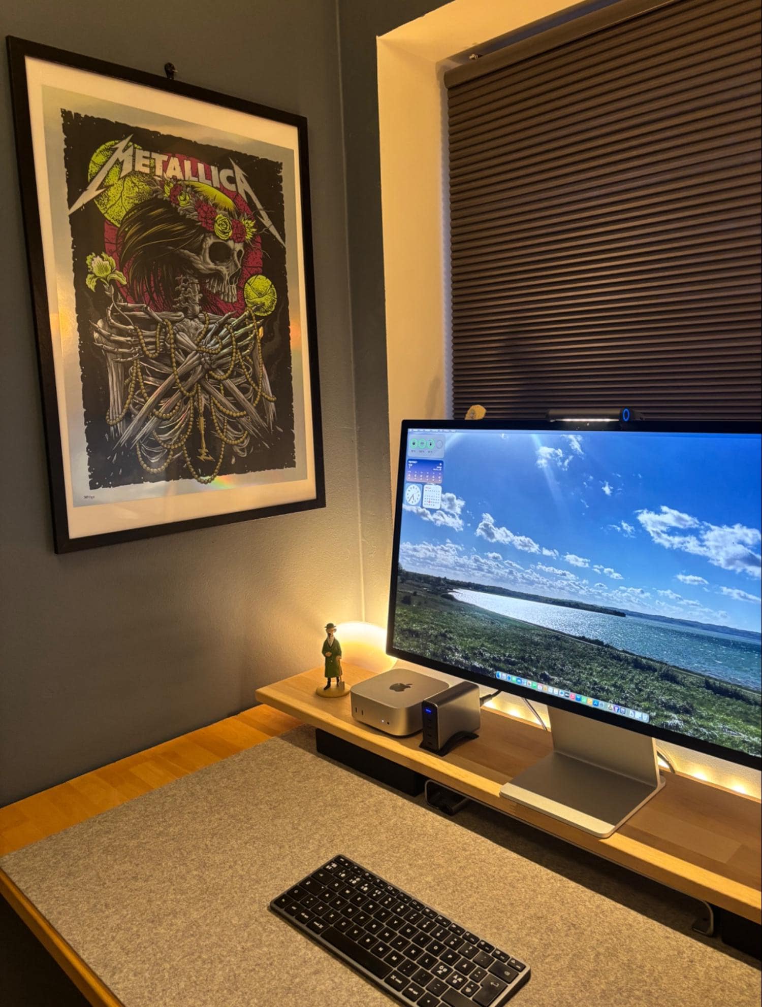 A home office desk with an Apple Studio Display, Mac Mini M4, Satechi keyboard, and a desk mat on a wooden desk with a wall shelf riser