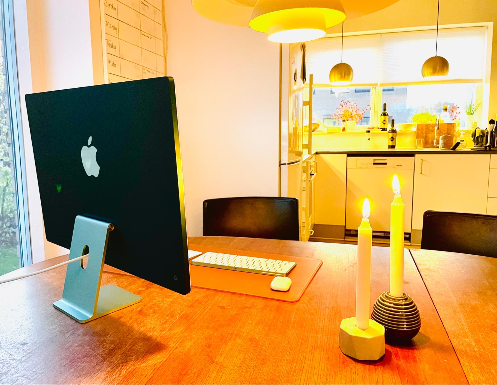 A small home workspace setup with an Apple desktop computer and wireless keyboard on a wooden dining table in a bright kitchen