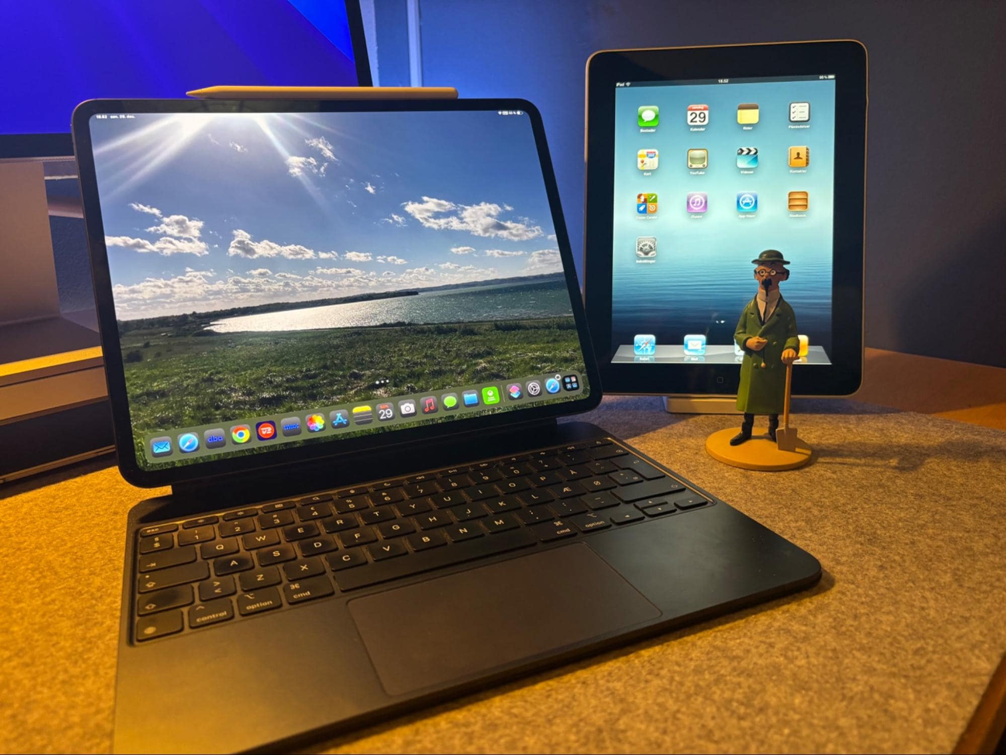 A workspace setup with an iPad Pro M4 13″ and Apple Pencil Pro on a Magic Keyboard next to an older iPad model and a small figurine, all on a felt desk mat