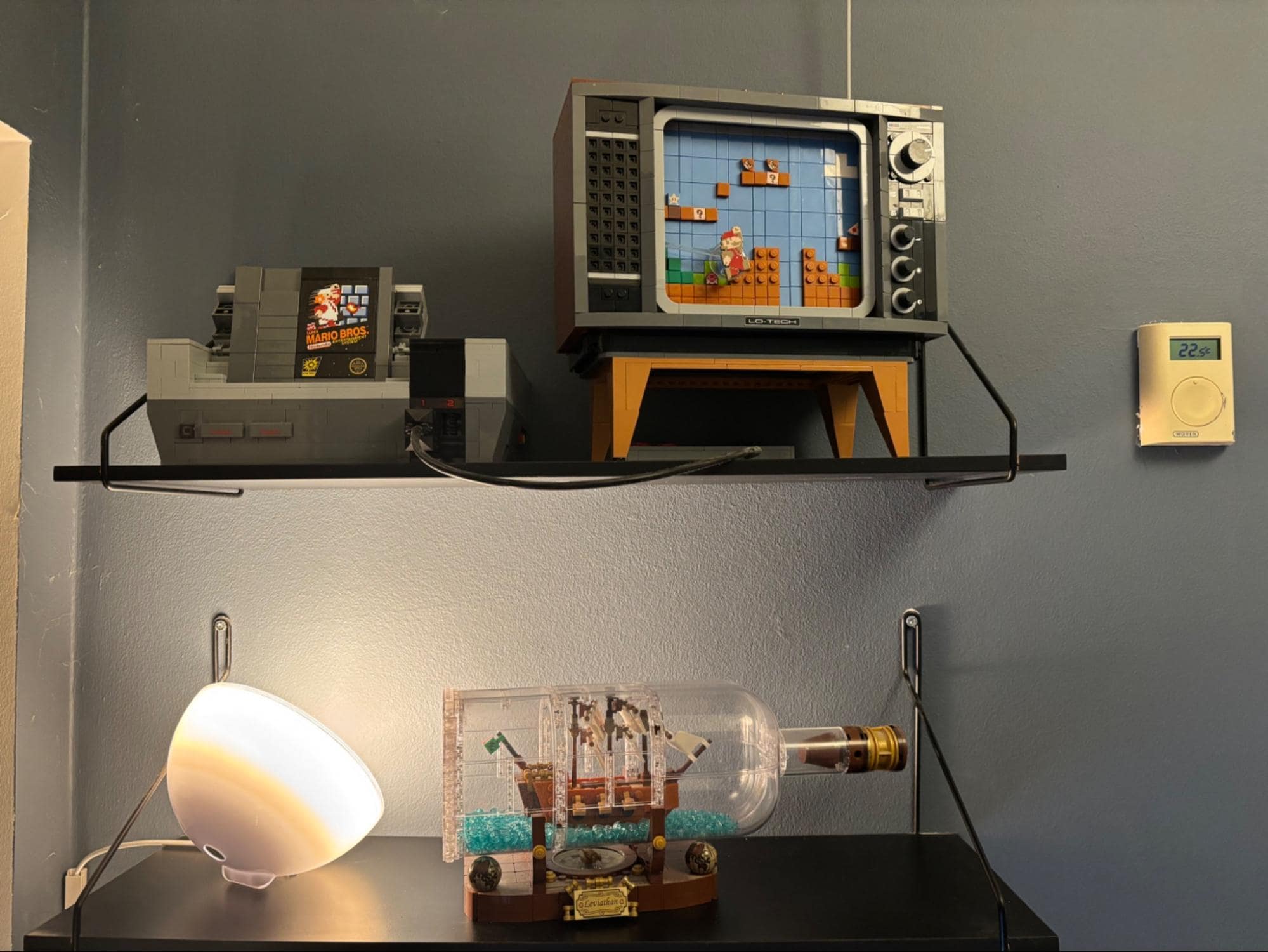 A shelf display with LEGO models of a retro TV showing Super Mario Bros., an NES console, and a ship in a bottle, with a wall-mounted thermostat showing 22°C