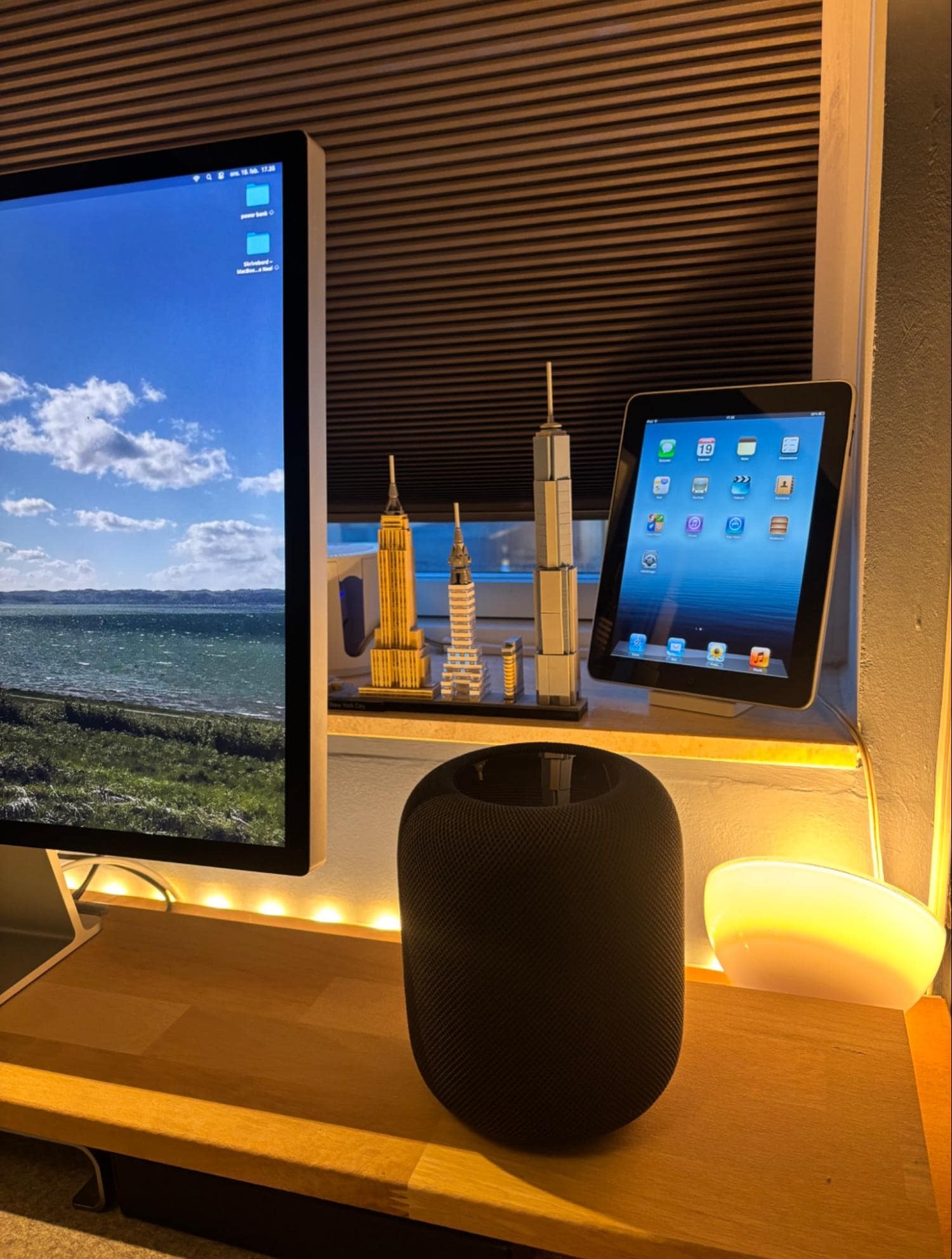 A home office corner with an Apple HomePod 2nd Gen, iPad Pro, and model skyscrapers on a wooden desk shelf next to an Apple Studio Display